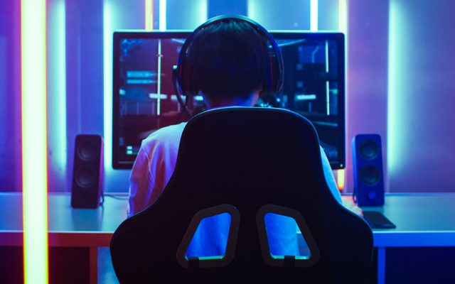 Back View Shot of the Beautiful Professional Gamer Girl Putting on Headset and Starts Playing Online Video Game on Her Personal Computer. Cute Casual Geek Girl. Room Lit by Neon Lamps in Retro Arcade Style.
